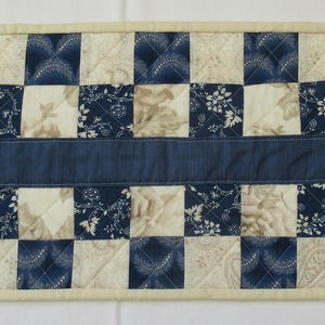 May include: A rectangular quilted table runner with a checkerboard design in navy blue and cream squares, some with floral patterns. A navy band runs down the centre. The runner is edged with cream fabric.