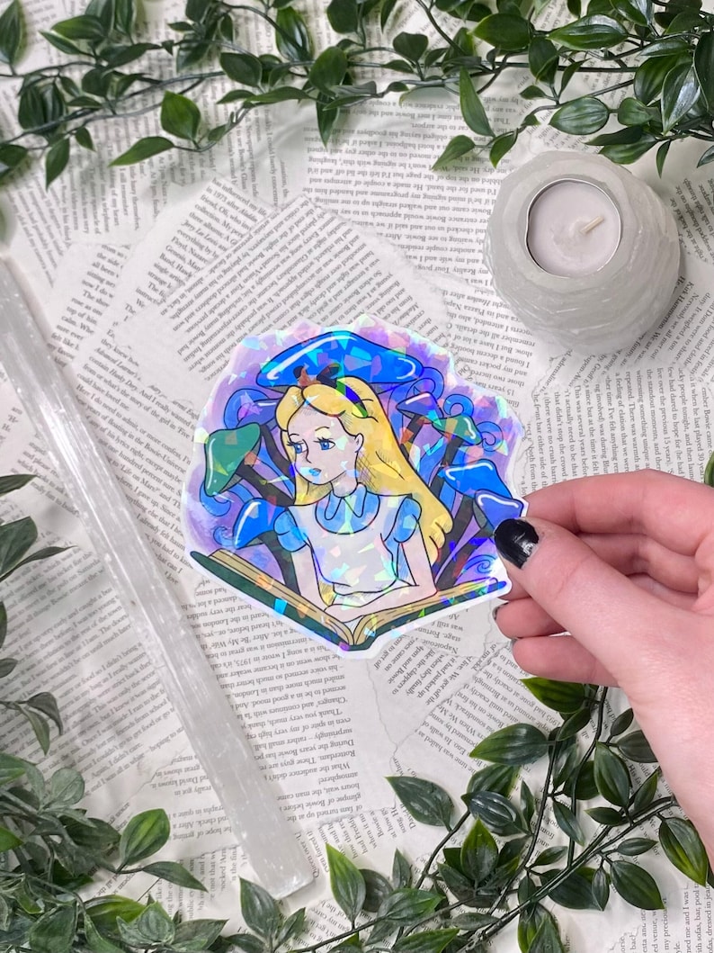 Puede incluir: Una pegatina hologr&aacute;fica con Alicia en el Pa&iacute;s de las Maravillas sentada con un libro en un campo de setas azules y moradas. La pegatina tiene un acabado iridiscente arco&iacute;ris.