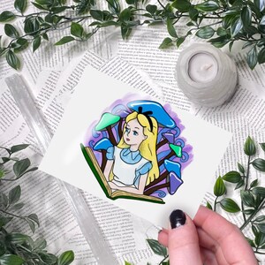 Puede incluir: Una ilustraci&oacute;n de acuarela de Alicia en el Pa&iacute;s de las Maravillas leyendo un libro en un bosque de setas moradas y azules. La ilustraci&oacute;n est&aacute; sobre un fondo blanco.