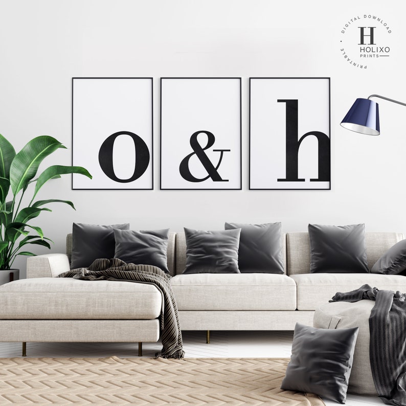 May include: Three black and white framed prints with the letters "o", "&", and "h" in a bold font. The prints are hanging on a white wall above a light gray sectional sofa with gray throw pillows.