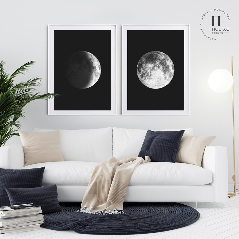 May include: Two black and white framed prints of the moon in different phases. The prints are hanging on a white wall above a white couch with beige and dark blue throw pillows and a beige throw blanket.