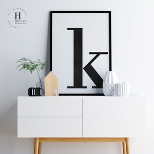 May include: A black and white printable wall art print featuring the letter "k" in a bold, modern font. The print is framed in a black frame and is displayed on a white dresser with a wooden top.
