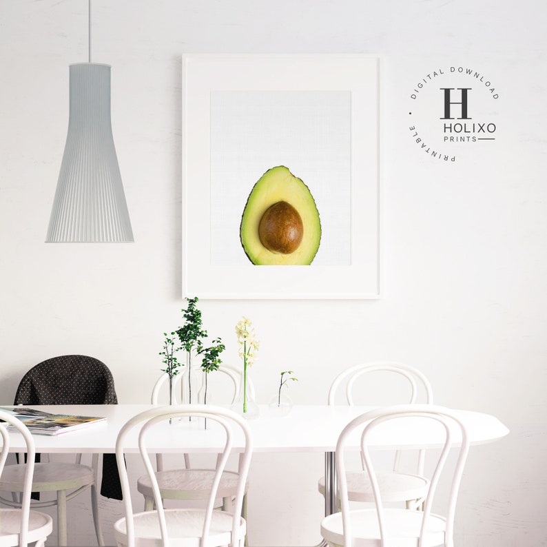 May include: A white framed print of a halved avocado with a brown pit. The print is hanging on a white wall in a dining room with a white table and white chairs. There are small potted plants on the table.