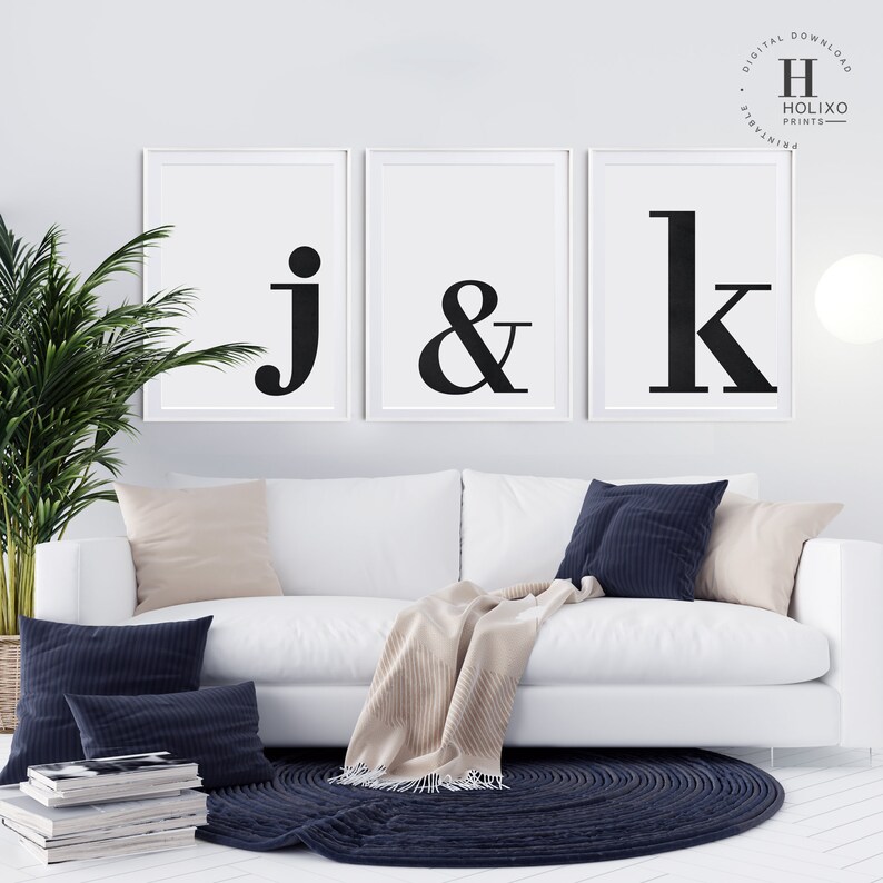 May include: Three black and white framed prints with the letters 'j', '&', and 'k' in a bold, modern font. The prints are hanging on a white wall above a white couch with beige and navy blue throw pillows.