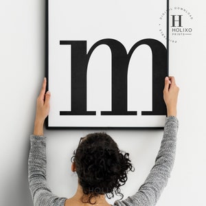 May include: A black and white printable wall art print featuring the letter "m" in a bold, sans-serif font. The print is framed in a black frame and is being hung on a white wall.