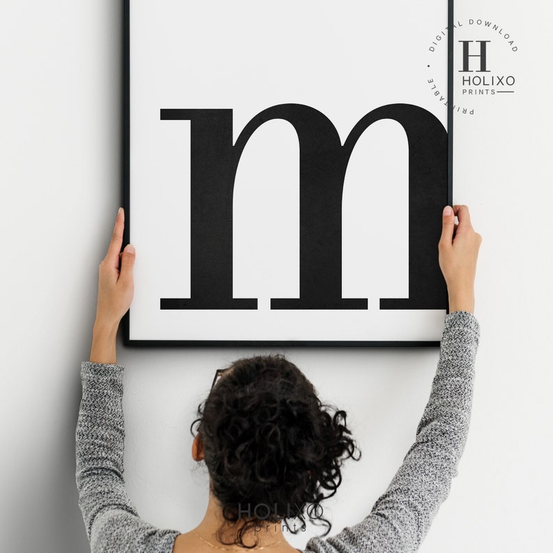 May include: A black and white printable wall art print featuring the letter "m" in a bold, sans-serif font. The print is framed in a black frame and is being hung on a white wall.
