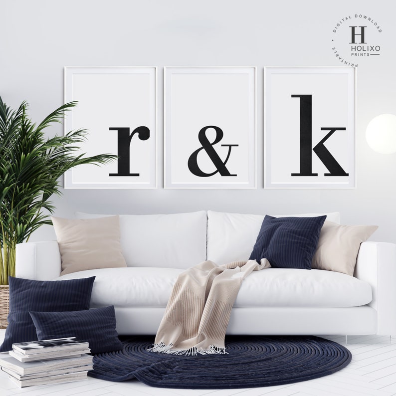 May include: Three black and white framed prints with the letters "r", "&", and "k" in a bold, sans-serif font. The prints are hanging on a white wall above a white couch with beige and navy blue throw pillows.