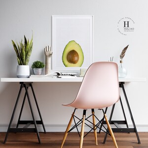 May include: A white framed print of a halved avocado with a white background. The print is on a white desk with a pink chair, a potted snake plant, a small potted plant, a silver hand sculpture, and a magazine.