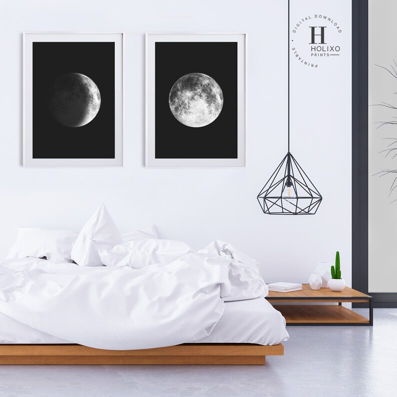 May include: Two black and white framed prints of the moon in different phases. The prints are hanging on a white wall above a white bed with a wooden headboard. A black geometric pendant light hangs above the bed.