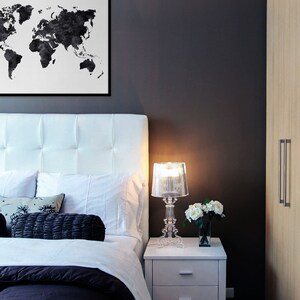 Puede incluir: Una cama tapizada en blanco con una impresi&oacute;n de mapa del mundo en blanco y negro colgada encima. Una mesita de noche blanca con dos cajones se encuentra junto a la cama, con una l&aacute;mpara de cristal transparente y un jarr&oacute;n de flores blancas en la parte superior.