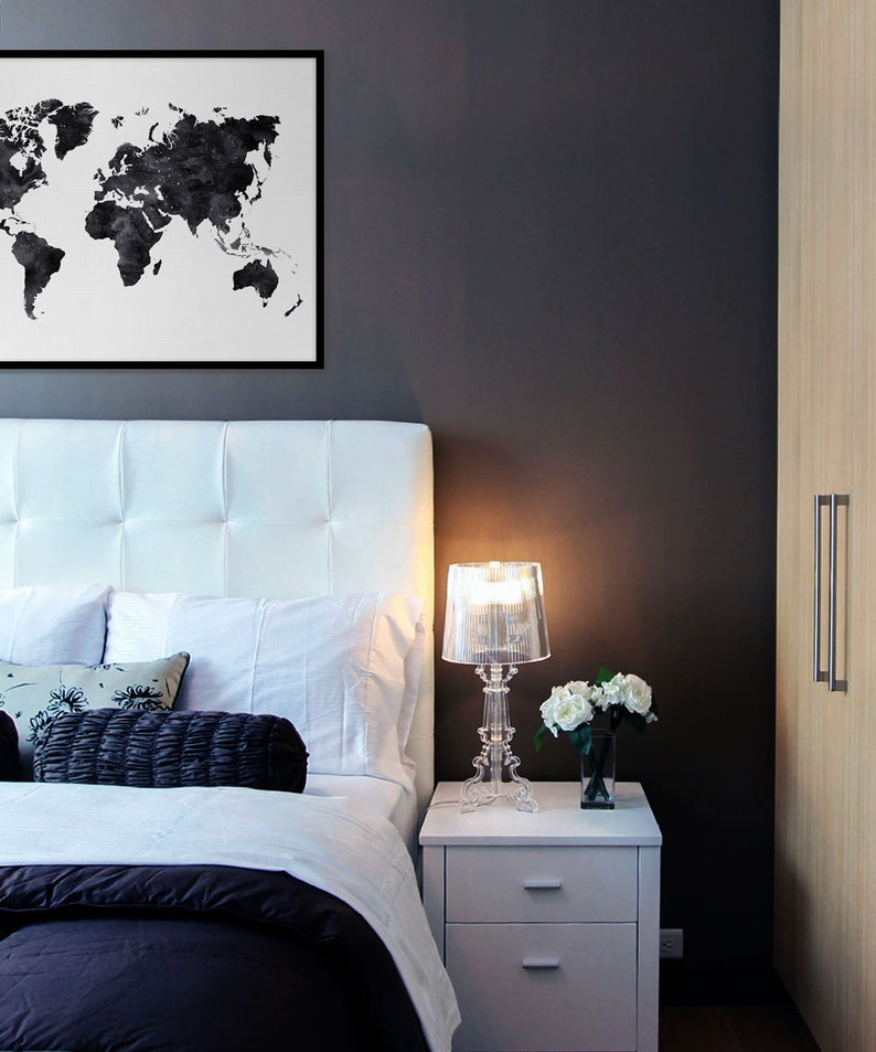 Puede incluir: Una cama tapizada en blanco con una impresi&oacute;n de mapa del mundo en blanco y negro colgada encima. Una mesita de noche blanca con dos cajones se encuentra junto a la cama, con una l&aacute;mpara de cristal transparente y un jarr&oacute;n de flores blancas en la parte superior.