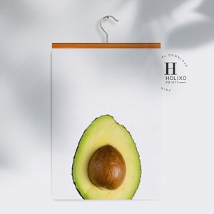 May include: A print of a halved avocado with a brown pit on a white background. The print is hanging on a wooden hanger with a metal hook. The print has a small logo in the top right corner that says "HOLIXO PRINTS".