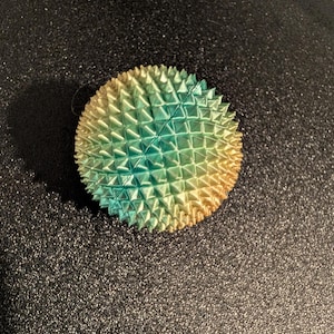 May include: A round, spiky object with a gradient of colors, transitioning from yellow to teal and blue. The object has a textured surface with numerous small, pointed protrusions, resembling a sea urchin or a stylized fruit.