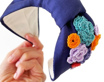 May include: A navy blue headband with a knotted design and white lining. It features crocheted flowers in orange, teal, purple, and red. The accessory is held by a hand, showcasing the colourful floral embellishments.