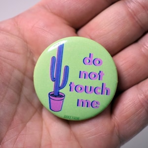 May include: A round, light green button with a cactus illustration and the words "do not touch me" in purple. The cactus is blue with a pink pot. The button is held in a hand.