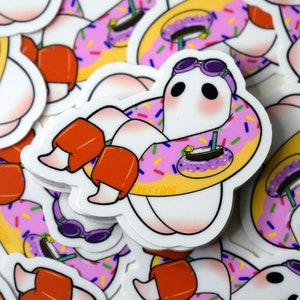 May include: A white ghost wearing purple goggles and orange arm floaties floats in a pink and yellow donut pool float with purple sprinkles. The ghost is holding a purple and yellow drink with a straw.