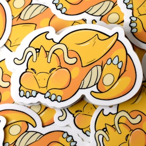K&ouml;nnte beinhalten: Ein Stapel gelber und wei&szlig;er Aufkleber mit einem schlafenden Cartoon-Drachen. Der Drache hat einen hellorangen Bauch und eine hellblaue Zunge.