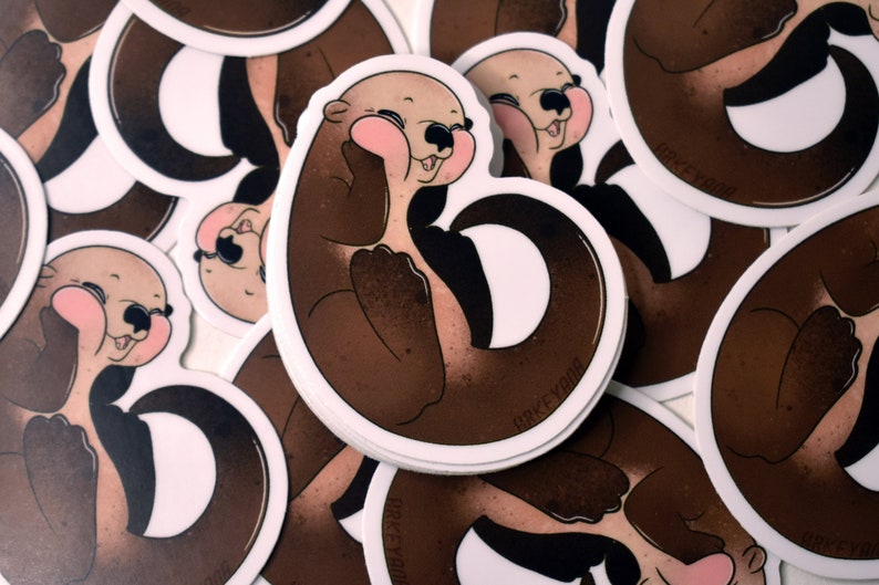 May include: A set of brown and white round stickers featuring a cartoon otter with a brown body and a white face. The otter is holding its paws up to its face and has a happy expression. The stickers have the text "BRKEYOOM" printed on them.