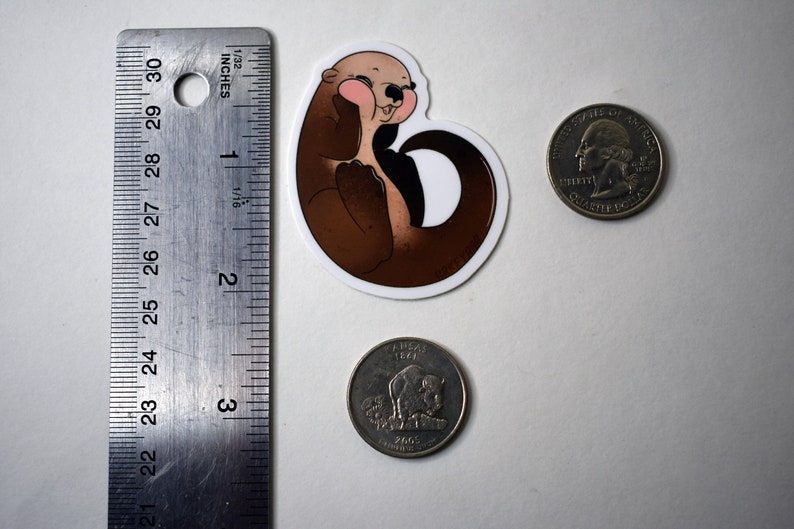 May include: A brown and white cartoon otter sticker with pink cheeks, smiling and holding its paws together. The sticker is about the size of a US quarter.