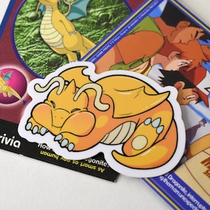 K&ouml;nnte beinhalten: Ein gelber und orangefarbener Cartoon-Drachen-Aufkleber mit wei&szlig;em Umriss. Der Drache ist zusammengerollt und schl&auml;ft. Der Aufkleber befindet sich auf einer blauen und wei&szlig;en Pokemon-Karte mit dem Text "MEWTWO STRIKES BACK".