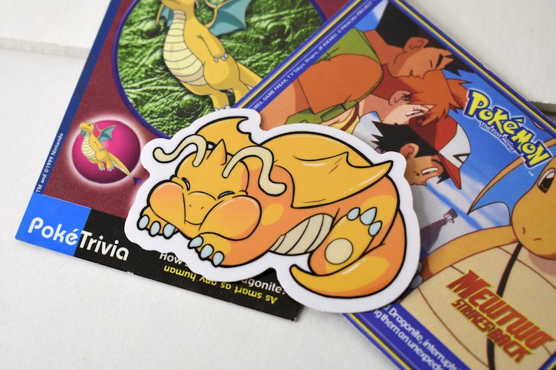 K&ouml;nnte beinhalten: Ein gelber und orangefarbener Cartoon-Drachen-Aufkleber mit wei&szlig;em Umriss. Der Drache ist zusammengerollt und schl&auml;ft. Der Aufkleber befindet sich auf einer blauen und wei&szlig;en Pokemon-Karte mit dem Text "MEWTWO STRIKES BACK".
