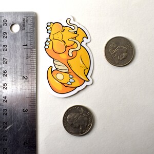 K&ouml;nnte beinhalten: Ein gelber und wei&szlig;er Cartoon-Drachen-Aufkleber, der sich schlafend zusammengerollt hat. Der Aufkleber ist etwa 5 cm breit und befindet sich neben einer 20-Cent-M&uuml;nze zum Gr&ouml;&szlig;envergleich.