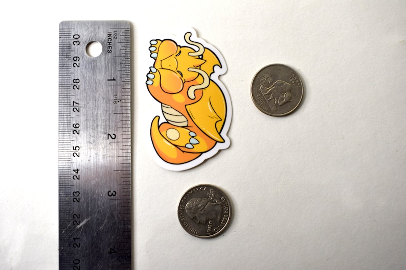 K&ouml;nnte beinhalten: Ein gelber und wei&szlig;er Cartoon-Drachen-Aufkleber, der sich schlafend zusammengerollt hat. Der Aufkleber ist etwa 5 cm breit und befindet sich neben einer 20-Cent-M&uuml;nze zum Gr&ouml;&szlig;envergleich.