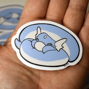 May include: A blue and white sticker featuring a cartoon character curled up in a ball, sleeping. The character has large ears and a white belly. The sticker is oval-shaped and has a white border.