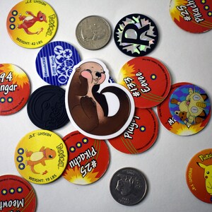 May include: A collection of round, colorful Pokemon trading card game tokens featuring various Pokemon characters, including Pikachu, Eevee, Gengar, and Meowth. A brown and white otter sticker is in the center of the image.