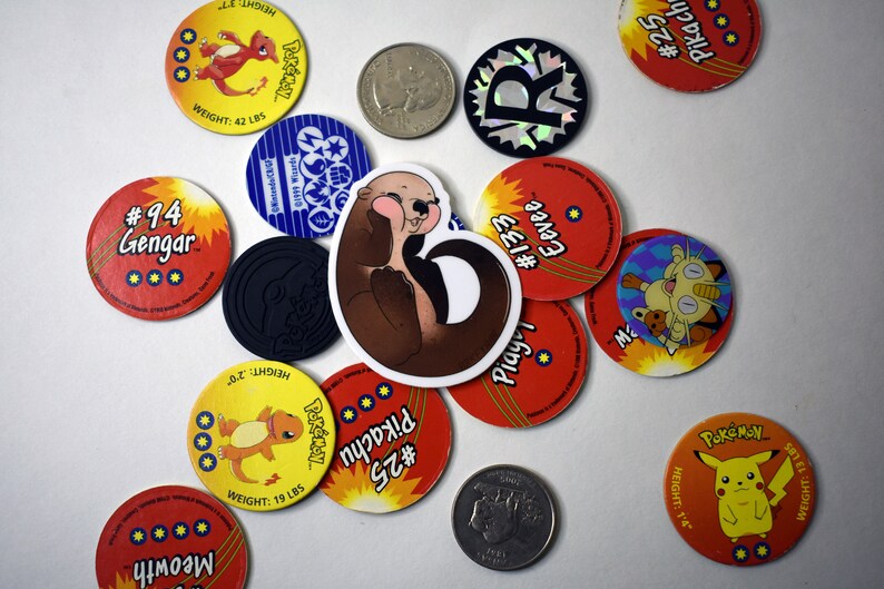 May include: A collection of round, colorful Pokemon trading card game tokens featuring various Pokemon characters, including Pikachu, Eevee, Gengar, and Meowth. A brown and white otter sticker is in the center of the image.