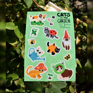 May include: A sheet of stickers featuring cats in a garden setting. The stickers are on a green background with a white border. The stickers feature cats wearing hats, holding gardening tools, and interacting with garden elements like ladybugs, mushrooms, and pumpkins. The text "Cats in the Garden" is at the top of the sheet.