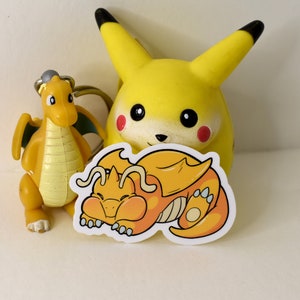 K&ouml;nnte beinhalten: Ein gelber Cartoon-Drachen-Aufkleber mit wei&szlig;em Rand. Der Drache schl&auml;ft mit dem Kopf auf seiner Pfote. Der Aufkleber befindet sich auf einer wei&szlig;en Oberfl&auml;che neben einem gelben Cartoon-Pikachu-Spielzeug.