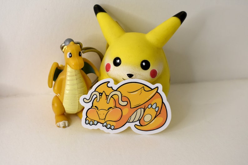 K&ouml;nnte beinhalten: Ein gelber Cartoon-Drachen-Aufkleber mit wei&szlig;em Rand. Der Drache schl&auml;ft mit dem Kopf auf seiner Pfote. Der Aufkleber befindet sich auf einer wei&szlig;en Oberfl&auml;che neben einem gelben Cartoon-Pikachu-Spielzeug.