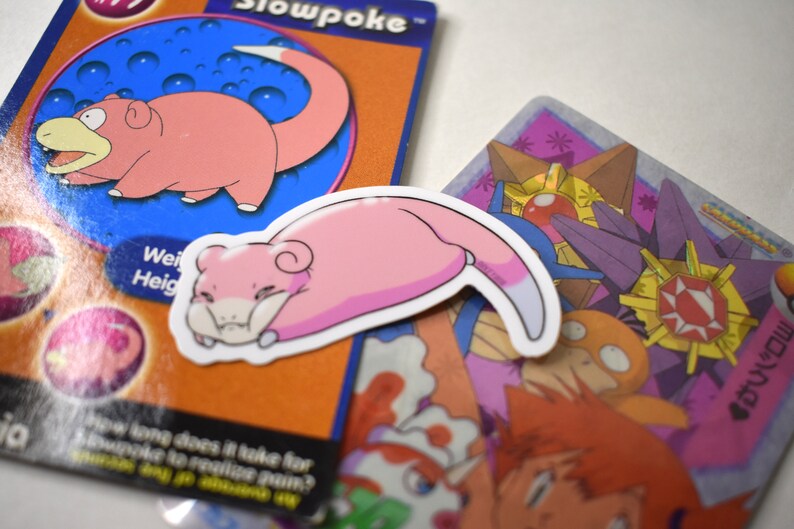 Puede incluir: Una pegatina rosa de Slowpoke, un Pok&eacute;mon, con un contorno blanco. La pegatina muestra a Slowpoke durmiendo de lado con la cola enroscada alrededor de su cuerpo. La pegatina est&aacute; encima de una carta de intercambio de Pok&eacute;mon con un fondo morado y dorado y una gema roja en el centro.