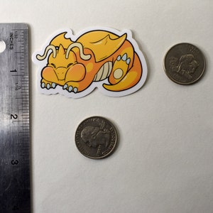 K&ouml;nnte beinhalten: Ein gelber und orangefarbener Cartoon-Drachen-Aufkleber mit wei&szlig;em Umriss. Der Drache ist zusammengerollt und schl&auml;ft. Der Aufkleber ist etwa so gro&szlig; wie eine deutsche 20-Cent-M&uuml;nze.