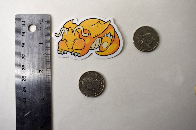 K&ouml;nnte beinhalten: Ein gelber und orangefarbener Cartoon-Drachen-Aufkleber mit wei&szlig;em Umriss. Der Drache ist zusammengerollt und schl&auml;ft. Der Aufkleber ist etwa so gro&szlig; wie eine deutsche 20-Cent-M&uuml;nze.