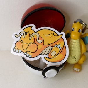 K&ouml;nnte beinhalten: Ein gelber und orangefarbener Cartoon-Drachen-Aufkleber mit wei&szlig;em Umriss, der in einem roten und wei&szlig;en Pok&eacute;ball schl&auml;ft.
