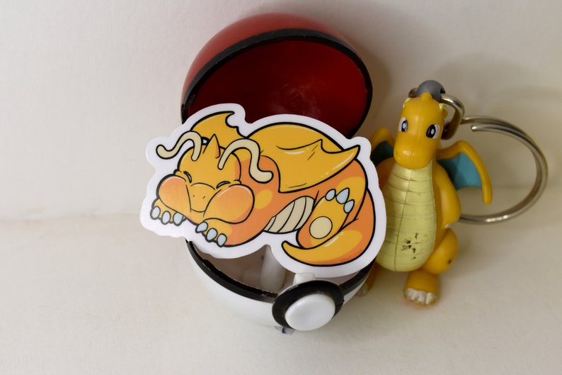 K&ouml;nnte beinhalten: Ein gelber und orangefarbener Cartoon-Drachen-Aufkleber mit wei&szlig;em Umriss, der in einem roten und wei&szlig;en Pok&eacute;ball schl&auml;ft.