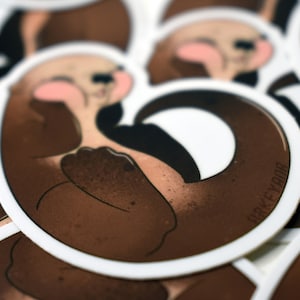May include: Close-up of a stack of brown and white round stickers featuring a cartoon otter with a curled tail. The otter is brown with a white belly and a pink nose. The text "ORKEYBOO" is printed on the sticker.