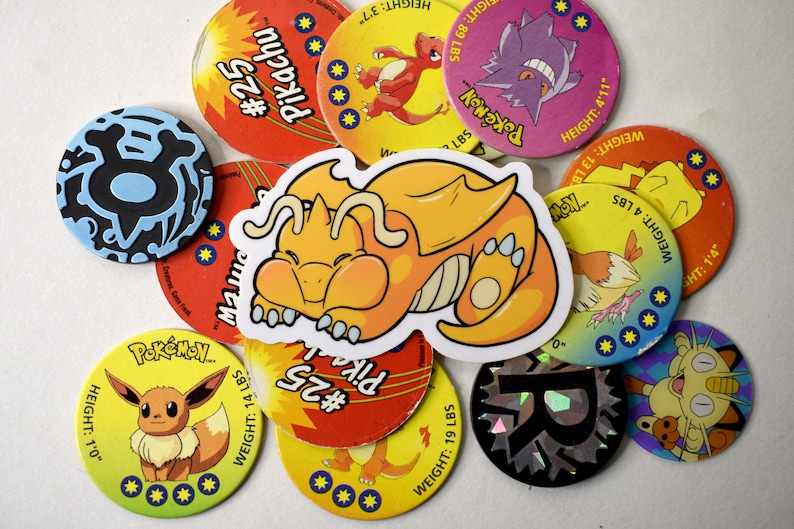 K&ouml;nnte beinhalten: Eine Sammlung von bunten Pok&eacute;mon-Sammelkarten mit verschiedenen Charakteren, darunter Pikachu, Glurak und Bisasam. Die Karten sind rund und haben einen gelben Hintergrund mit schwarzem Text und Bildern. Die Karten liegen auf einem wei&szlig;en Untergrund in einem Stapel.