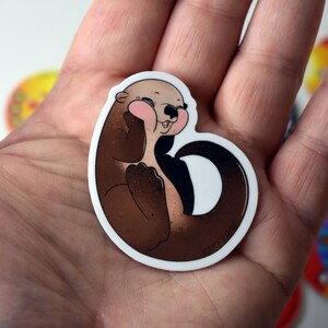 May include: A brown and white cartoon otter sticker with pink cheeks and a white background. The otter is curled up with its tail wrapped around its body.