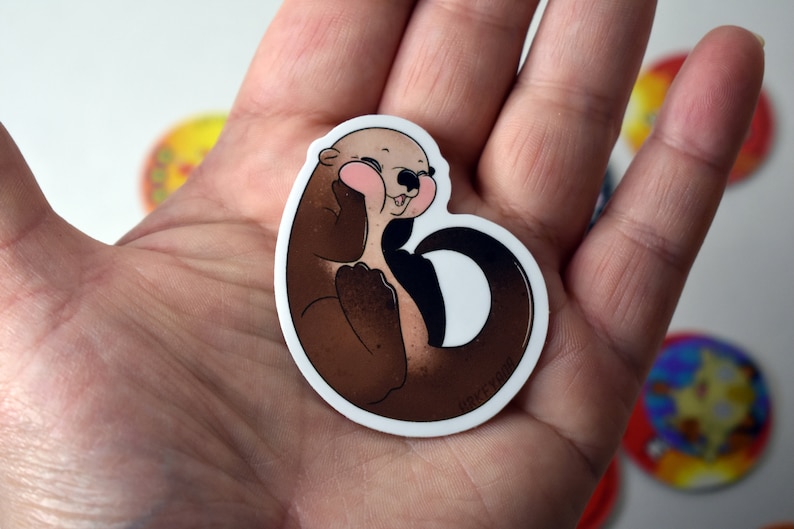 May include: A brown and white cartoon otter sticker with pink cheeks and a white background. The otter is curled up with its tail wrapped around its body.