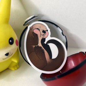 May include: A brown and white cartoon otter sticker with pink cheeks, inside a red and black Pokeball.
