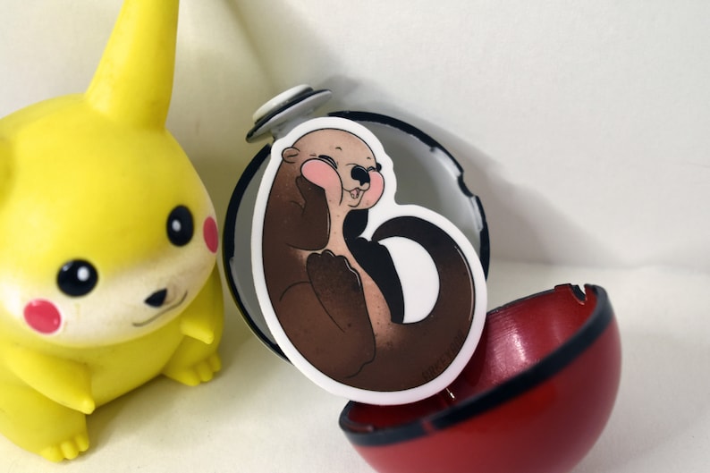 May include: A brown and white cartoon otter sticker with pink cheeks, inside a red and black Pokeball.