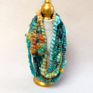 May include: A multi-strand infinity scarf featuring a blend of textured yarns in shades of teal, turquoise, emerald green, and golden yellow. The scarf incorporates metallic gold threads and small sequins throughout the various yarn weights.