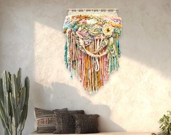 May include: A colorful, textured fiber art wall hanging. The piece features a variety of yarns in pastel shades of pink, yellow, green, and blue, with a cream-colored lace top and a wooden dowel for hanging. The design includes knotted and fringed elements.