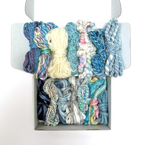 May include: A white cardboard box filled with an assortment of yarn skeins in various shades of blue, cream, white, and multi-colored pastel blends. The collection includes different textures, ranging from smooth plied yarn to thick, nubby, and variegated strands arranged in two horizontal rows.