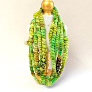 May include: A long, textured scarf featuring multiple strands of yarn in various shades of lime, emerald, and olive green. The yarn has a coiled, spiral appearance with varying thicknesses and fuzzy textures, draped over a mannequin torso.