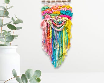 Puede incluir: Un vibrante tapiz de pared hecho a mano con un arcoíris de colores. La pieza de arte textil incluye hilos rosas, amarillos, naranjas, verdes y azules, con una varilla de madera en la parte superior para colgar. Una flor rosa está unida al frente.