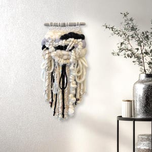 May include: A handcrafted fiber art wall hanging. The piece features a combination of white, black, and gray yarns and textures. A small, white flower is attached to the front. The wall hanging is mounted on a natural wooden dowel.